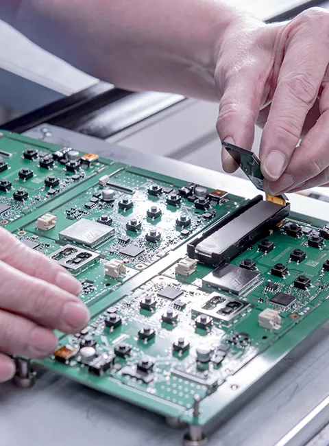 Hands working on a green circuit board with electronic components in a manufacturing environment.