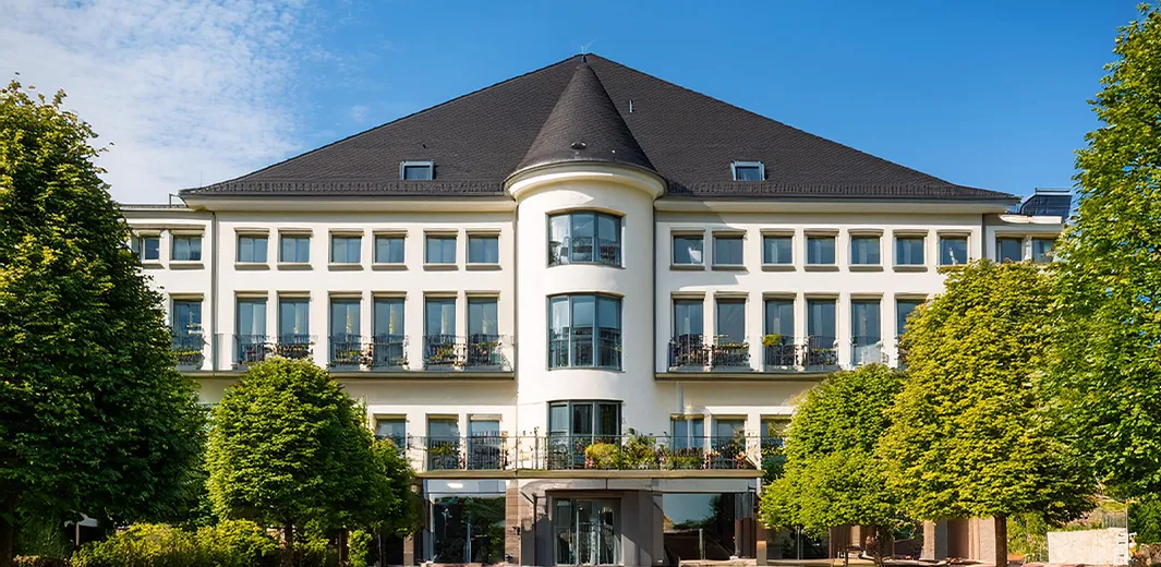 Elegantes, mehrstöckiges Gebäude im Stil einer Residenz oder eines Hotels, mit symmetrischer Fassade und gepflegter Baumallee.