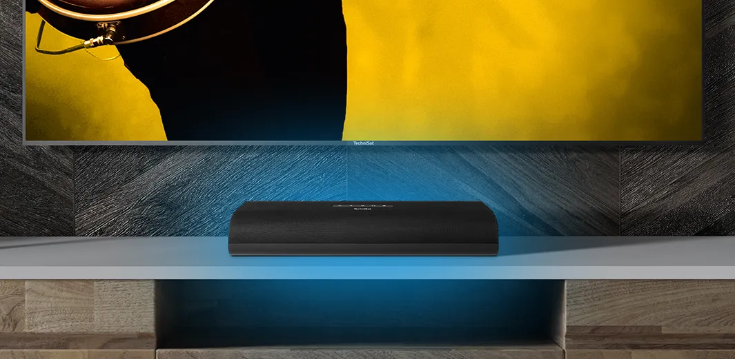 A TechniSat soundbar sits on a shelf in the living room in front of a TV, delivering excellent sound.