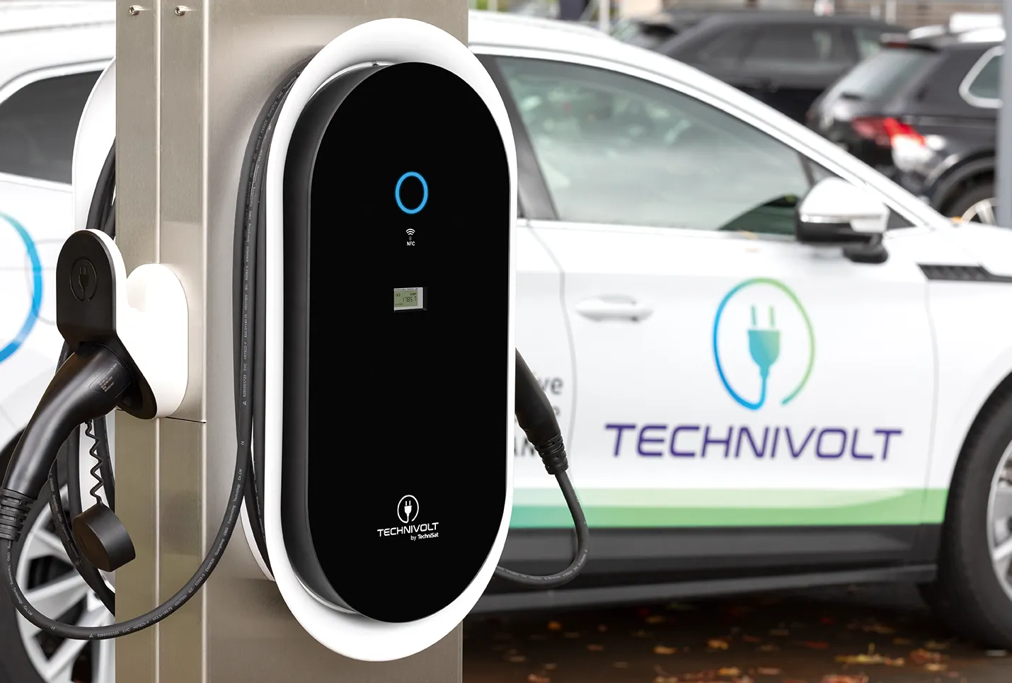 Nahaufnahme einer TECHNIVOLT-Wallbox auf einem Firmenparkplatz mit mehreren Elektroautos im Hintergrund.