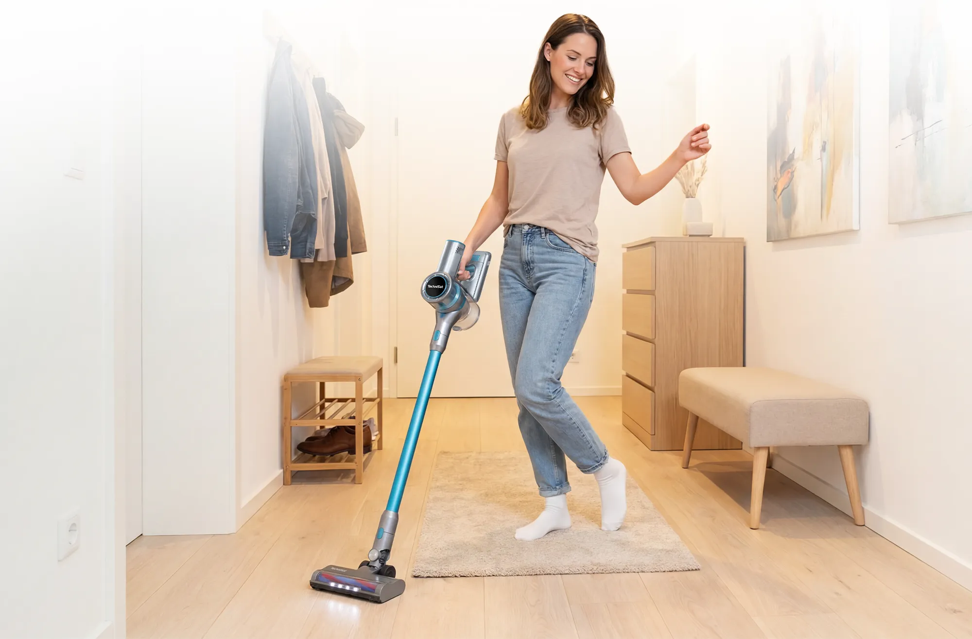 Eine lächelnde Frau saugt mit einem kabellosen Staubsauger einen Teppich in einem hellen Flur zuhause.