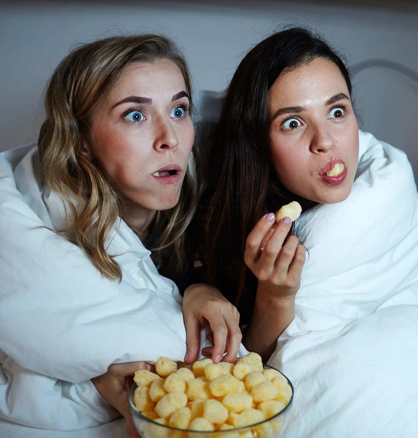 Bild von zwei Frauen, die gespannt und mit großen Augen vor einem Fernseher sitzen, in eine Bettdecke eingewickelt und dabei essen.