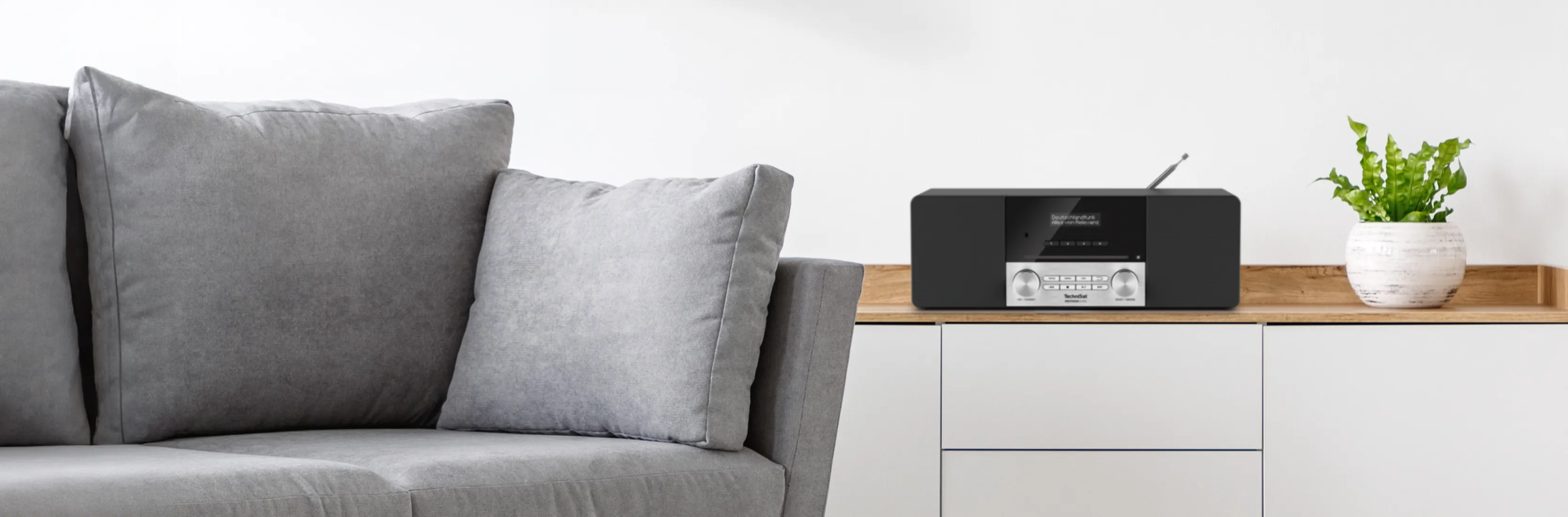Modernes Stereoradio von TechniSat (Modell DIGITRADIO 3) platziert auf einem Sideboard in aufgeräumtem Wohnzimmer. Ein Couch lädt zum entspannten Radiohören ein.